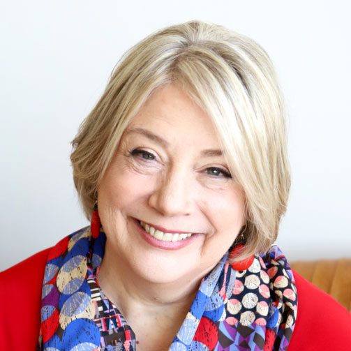 Smiling older woman with short, blonde hair, wearing a colorful patterned scarf and a red top, seated against a plain, light background.