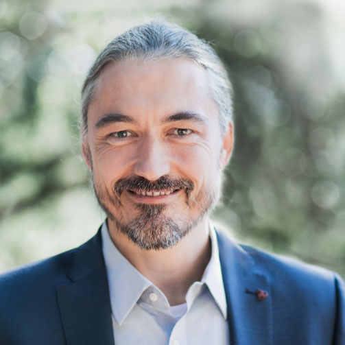 A man with long, gray hair tied back and a beard, wearing a suit jacket and light shirt, smiles outdoors with a blurred green background.