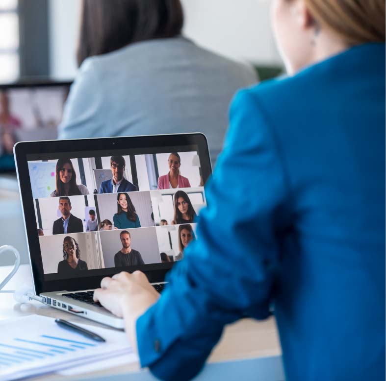 Person in a blue blazer participates in a video conference on a laptop, with multiple people visible in a grid layout on the screen.