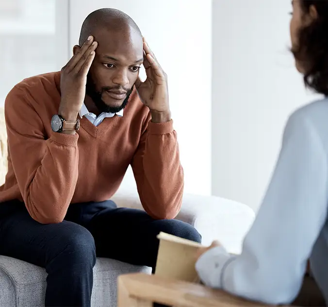 A man sits on a couch holding his head in his hands, appearing stressed, while talking to a therapist during a counseling session.