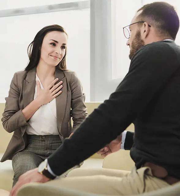Two people sit on a couch having a conversation; the woman has her hand on her chest and is smiling, while the man listens attentively.