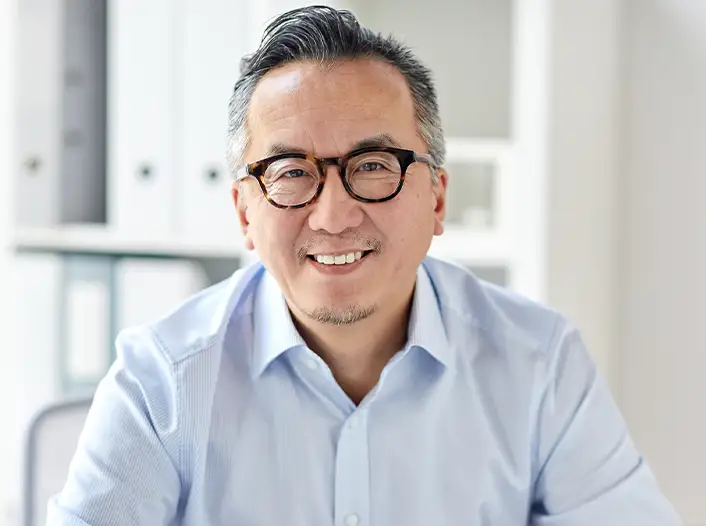 A man with glasses and gray hair, wearing a light blue collared shirt, is smiling while sitting indoors in a well-lit office setting.