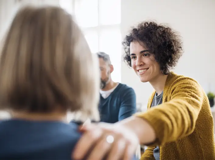 A woman with curly hair and a yellow sweater smiles and places her hand supportively on another person's shoulder during a group meeting.