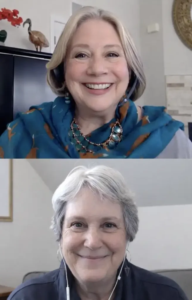 Two older women smiling during a video call, one wearing a blue scarf and necklace, the other with short gray hair and earphones.