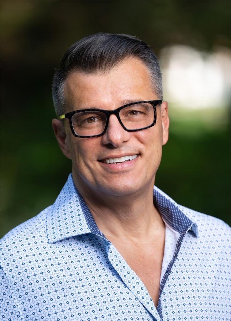 A man with short gray hair and glasses wearing a patterned button-up shirt smiles outdoors with a blurred green background.