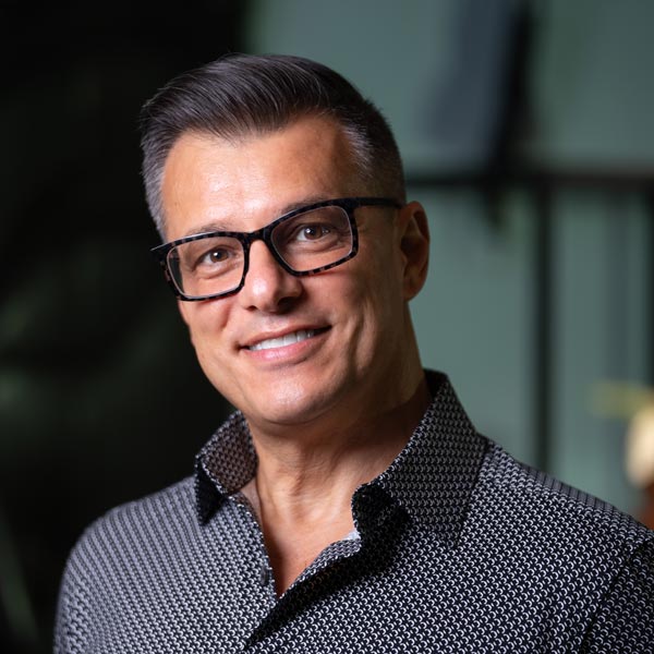 A man with short, dark hair and glasses smiles at the camera, wearing a patterned button-up shirt. The background is blurred.