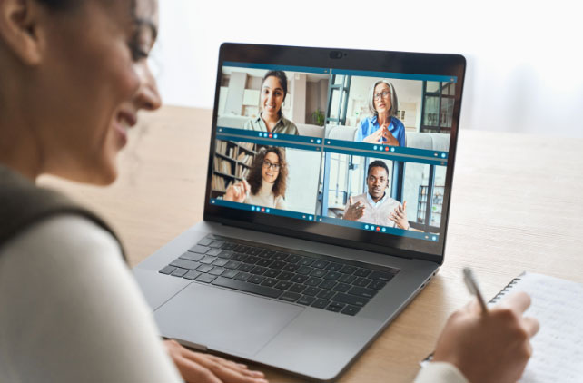 A person takes notes while participating in a video conference call with four people displayed on a laptop screen.