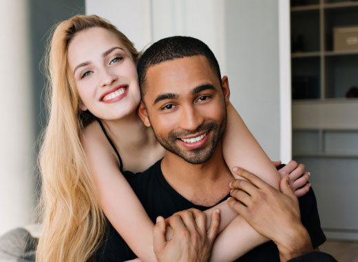 A woman with long blonde hair hugs a man from behind; both are smiling and looking at the camera in a modern indoor setting.