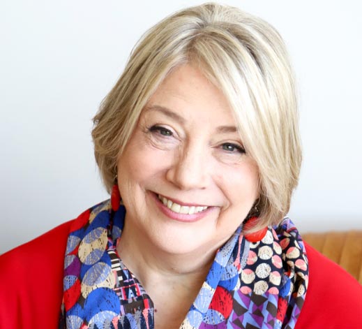 Smiling older woman with short gray hair, wearing a colorful patterned scarf and red top, pictured against a plain light background.