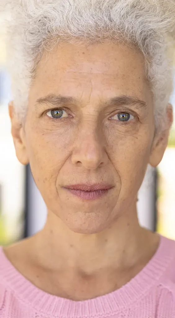 An older woman with short curly white hair and light-colored eyes looks directly at the camera. She is wearing a light pink sweater and has a neutral expression.