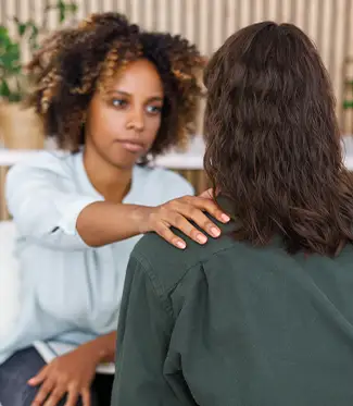 A woman places her hand on another person's shoulder, offering support during a conversation in an indoor setting.