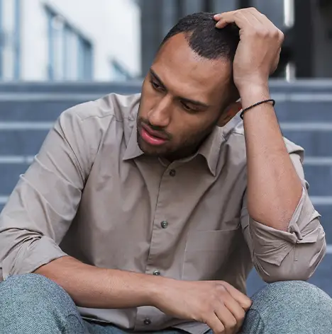 A man sits on outdoor steps, looking down with a concerned expression and holding his head with one hand.