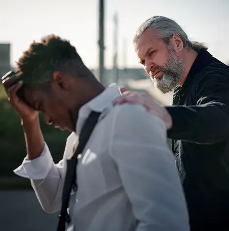 An older man comforts a younger man who is visibly upset, with his head down and hand on his forehead.