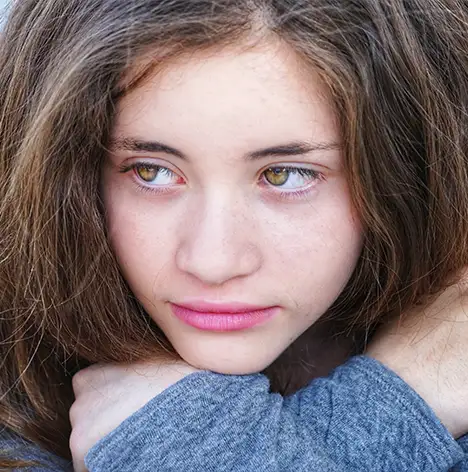Young person with long brown hair and hazel eyes rests their chin on crossed arms, gazing slightly downward with a neutral expression.