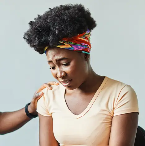 A woman with a colorful headband sits looking upset while another person places a comforting hand on her shoulder.