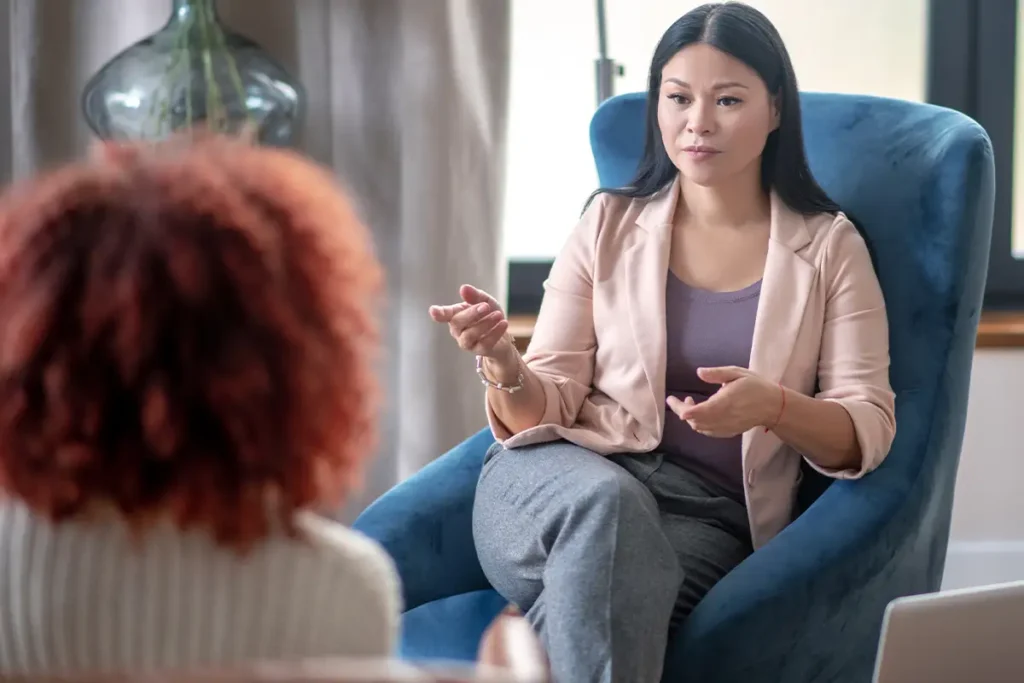 A woman in a blazer sits in a blue chair, gesturing as she speaks to another person with curly hair in a counseling or therapy session.