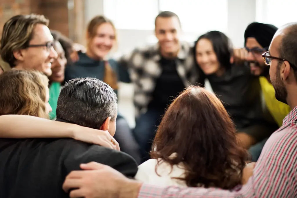 A group of people sit together in a close circle with arms around each other, smiling and appearing to enjoy each other's company.