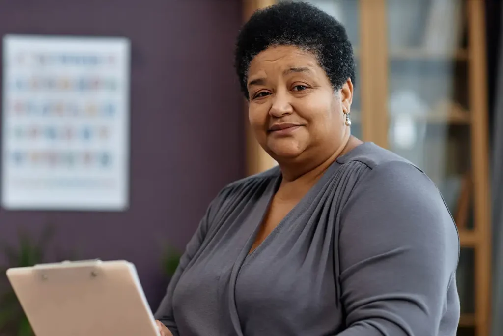 A person with short curly hair and a gray top holds a clipboard and looks at the camera, standing indoors with a blurred background.