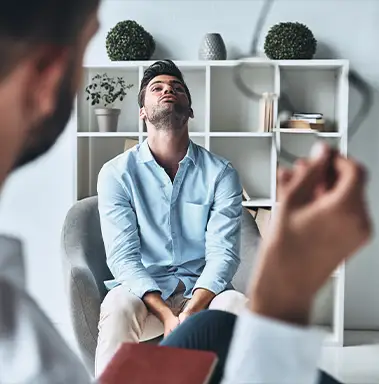 A man sits back in a chair, looking up with a frustrated expression, while another person in the foreground holds a notebook and pen during a conversation.