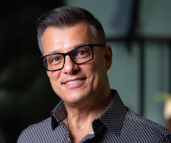 A man with short, neatly styled gray hair and glasses is smiling at the camera. He is wearing a patterned button-up shirt.