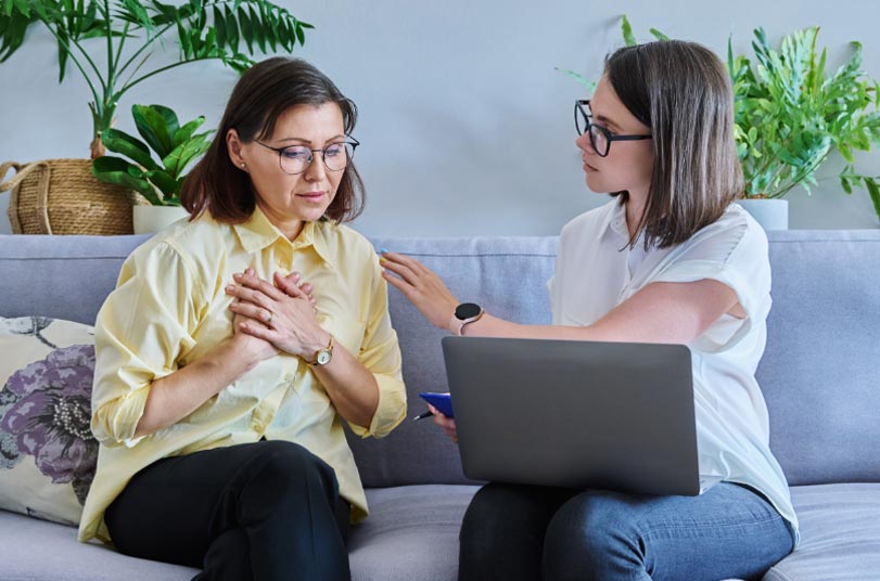 Two women sit on a couch; one holds her chest and looks concerned, while the other, with a laptop, offers comfort by touching her arm.