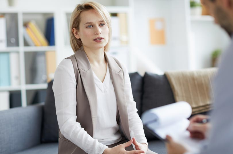 A woman sits on a couch talking to a person holding a notepad in a bright office setting, possibly during a counseling or therapy session.
