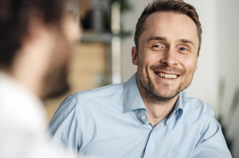 A man in a light blue shirt smiles while talking to another person in a blurred foreground, indoors.