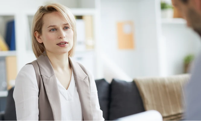 Woman with short blonde hair listens attentively to another person during a conversation in a bright, modern office setting.