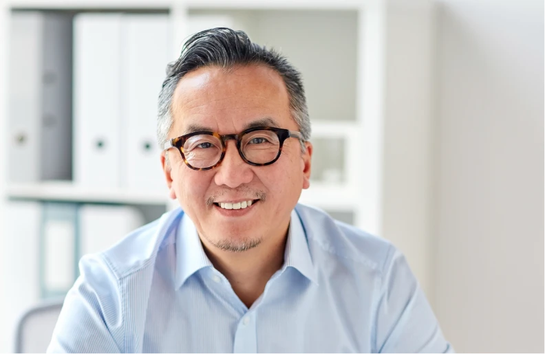A middle-aged man with glasses and gray hair smiles at the camera, wearing a light blue shirt and sitting in an office with shelves in the background.
