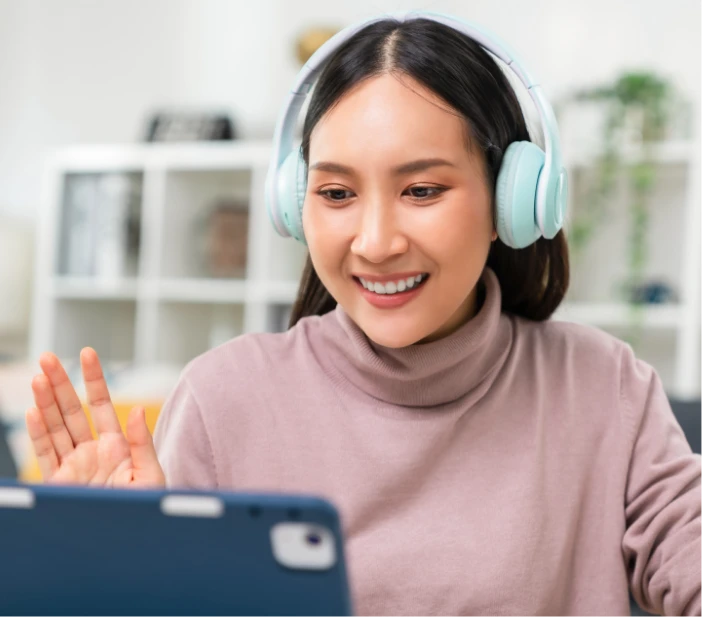 Woman wearing headphones smiles and waves at a tablet screen during a video call in a bright indoor setting.