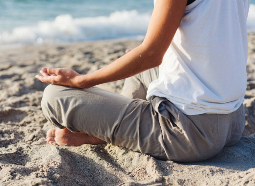 Person sitting cross-legged on the sand near the ocean, meditating with hands resting on knees.