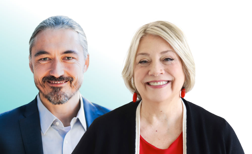 A man in a suit and a woman in a black sweater and red top stand side by side, smiling at the camera against a light gradient background.