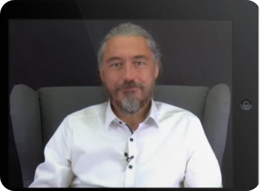 A man with gray hair and beard, wearing a white shirt, sits on a gray armchair and looks at the camera.