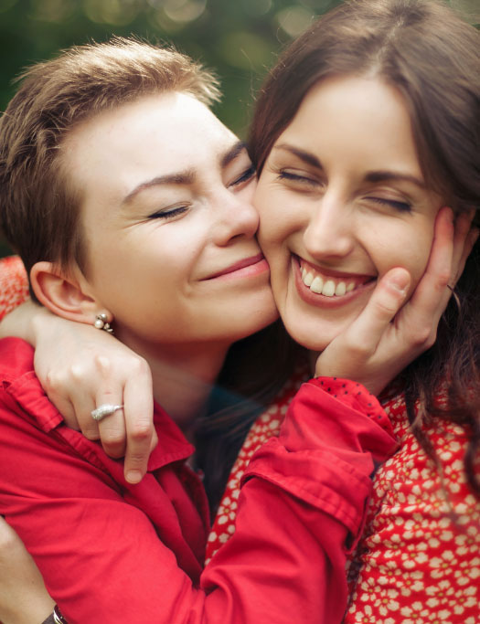 Two women wearing red clothing embrace and smile with eyes closed, one gently holding the other's face in an affectionate gesture, outdoors.