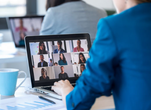 A person participates in a video conference call on a laptop, with multiple participants visible on the screen; a coffee mug and documents sit nearby.
